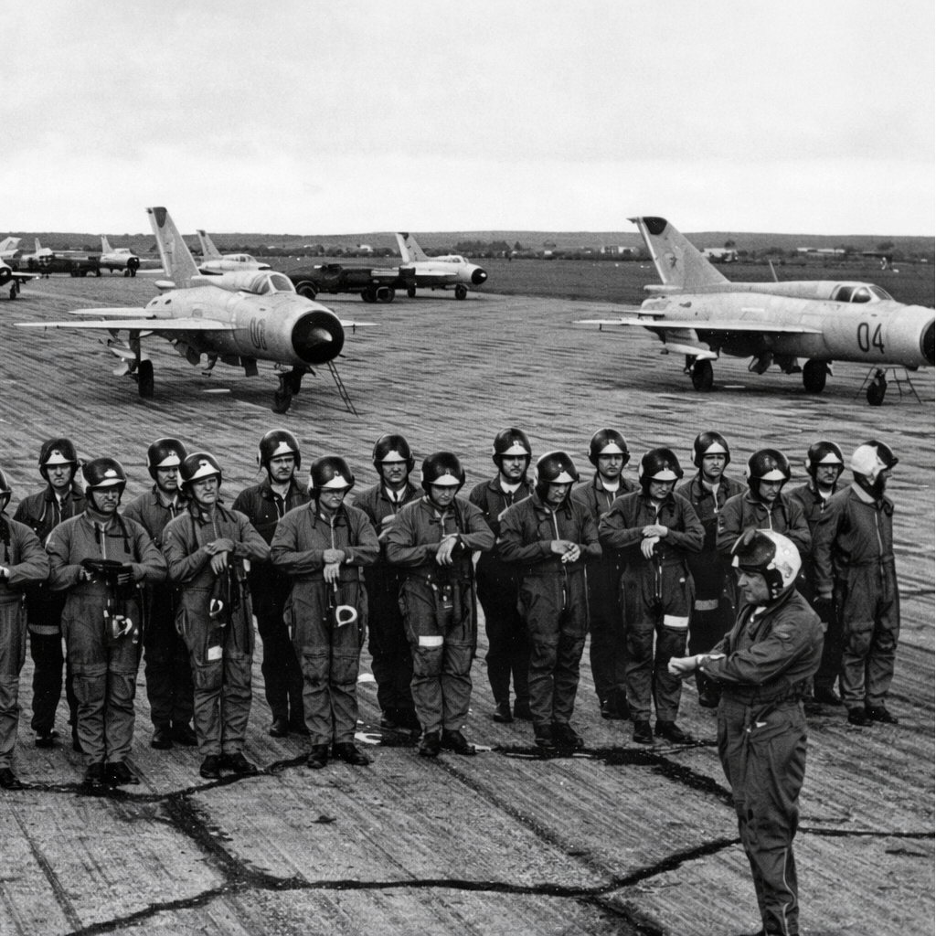 Group of people in uniform standing on an airfield with aircraft in the background, on Soviet hexagon tarmac